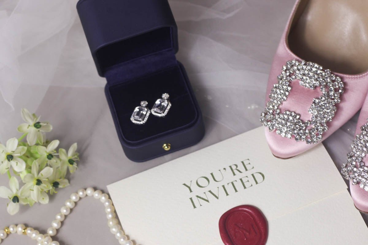 Pink Manolo Blanik Hangsili shoes with crystal embellishments, wedding invitation card, pearl necklace, and jewelry box with a personalized 18k gold diamond and sapphire earrings on a neutral background